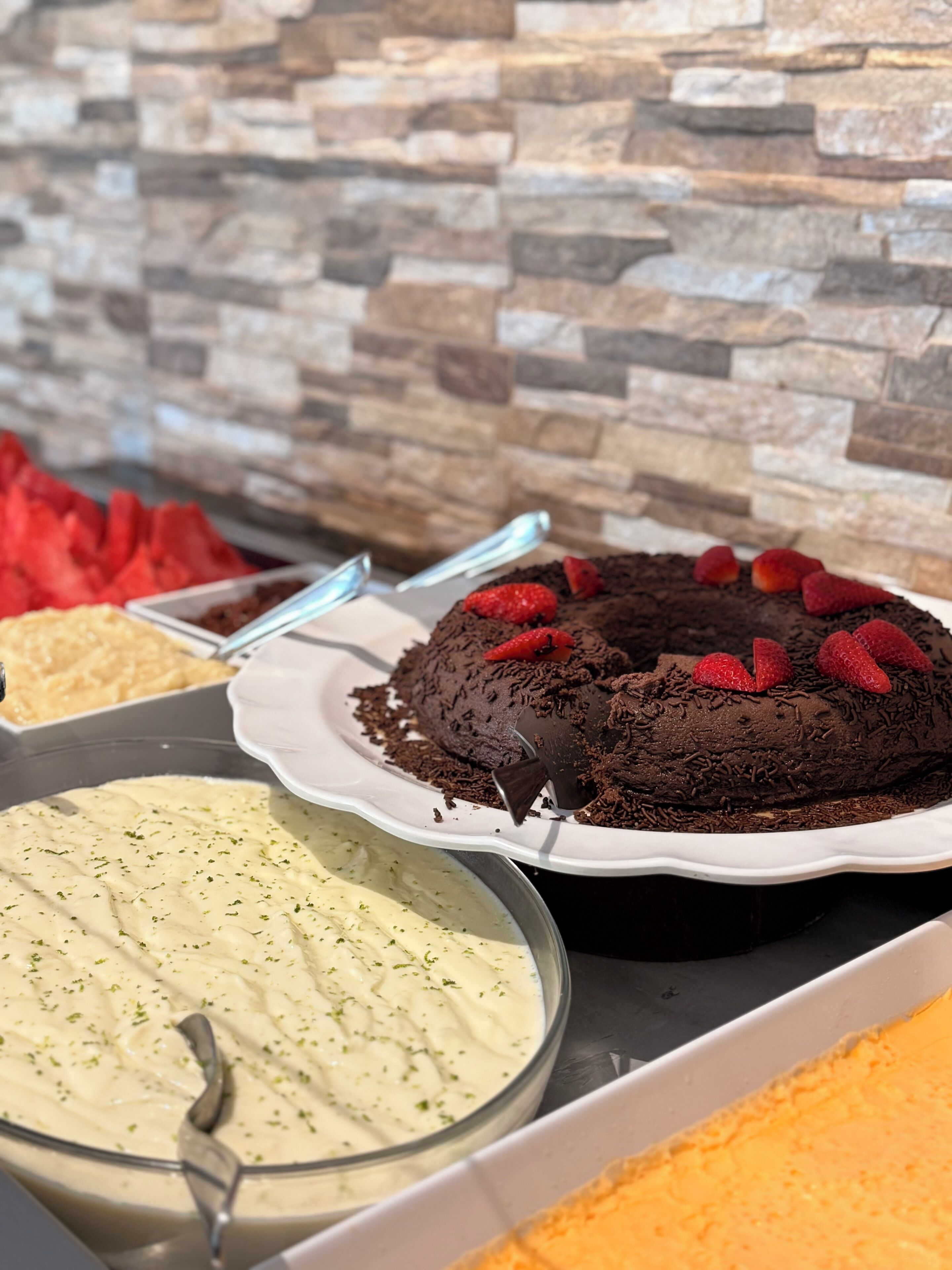 Um buffet de sobremesas com diversas opções, incluindo um bolo de chocolate coberto com morangos, uma tigela de molho verde e um recipiente com molho alaranjado.