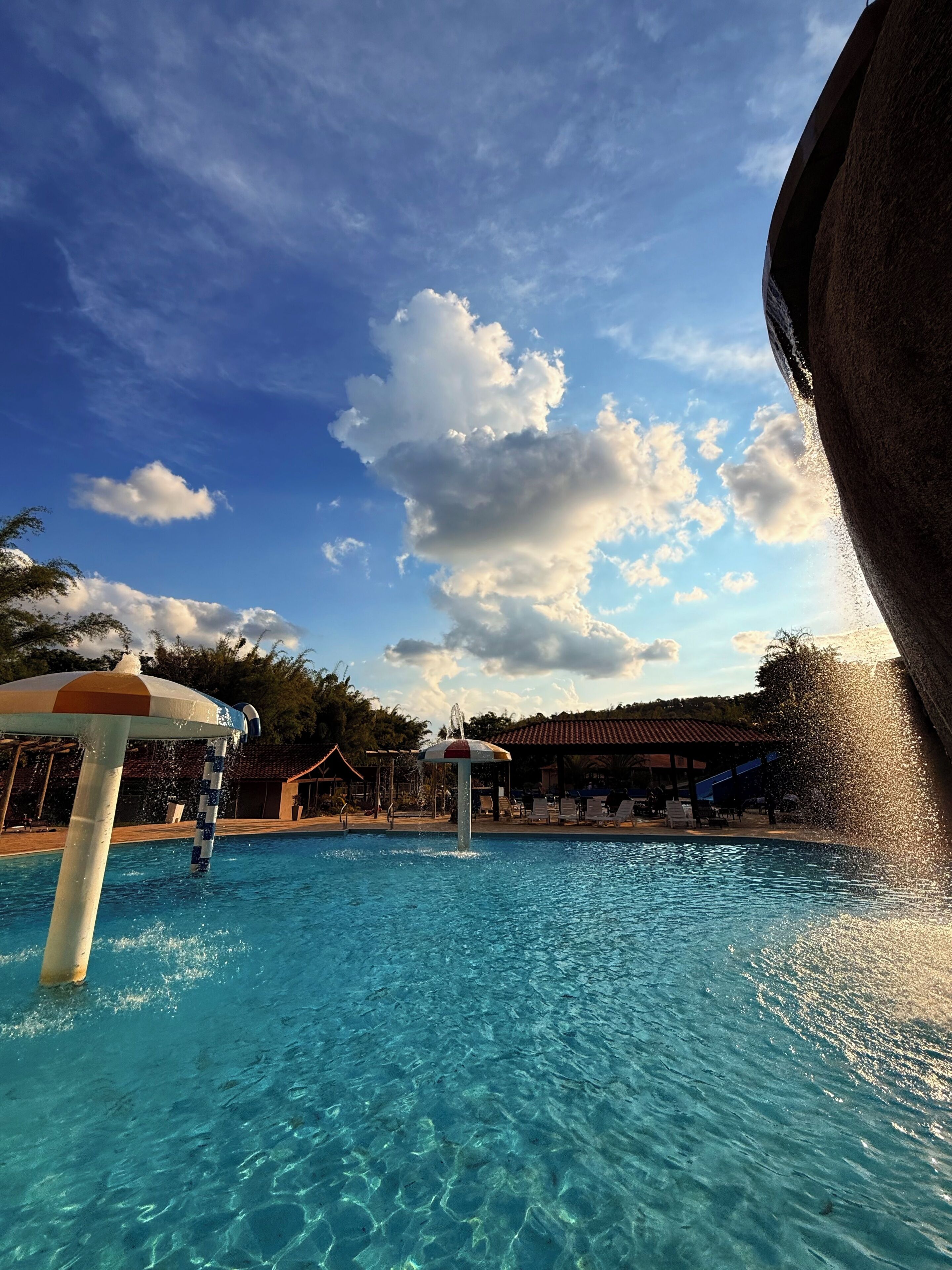 A clear blue pool with a water fountain, surrounded by lounge chairs and umbrellas, under a partly cloudy sky.