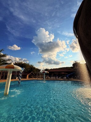 A clear blue pool with a water fountain, surrounded by lounge chairs and umbrellas, under a partly cloudy sky. - Hotel Campestre Atibaia (Atibaia)