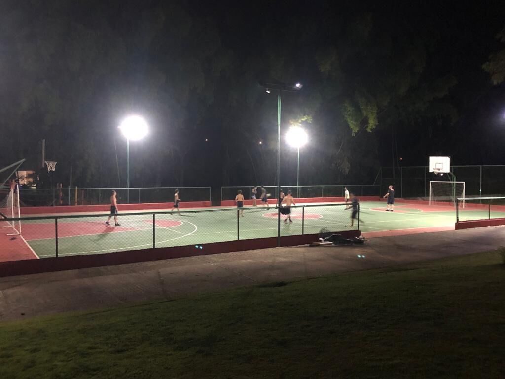 An outdoor basketball court at night with floodlights, surrounded by a fence and trees.