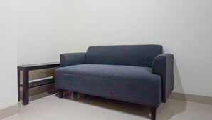 A corner room with a dark blue sofa, a small wooden side table, and a white wall. - Exquisite 1BR at Praxis Apartment (Surabaya)