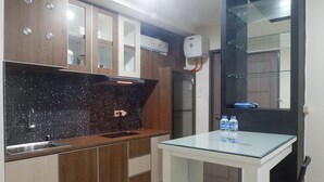 A modern kitchen with wooden cabinets, a black tiled backsplash, and a glass-top table with water bottles. - Exquisite 1BR at Praxis Apartment (Surabaya)