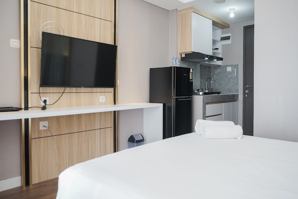 A modern hotel room with a large bed, a flat-screen TV mounted on a wooden cabinet, a built-in kitchen with a refrigerator and stove, and a small trash bin.