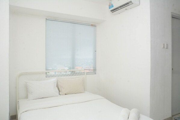 A small, white room with a single bed, a window with blinds, and an air conditioning unit on the wall. - Best and Strategic Studio at Bassura Apartment (Jakarta)