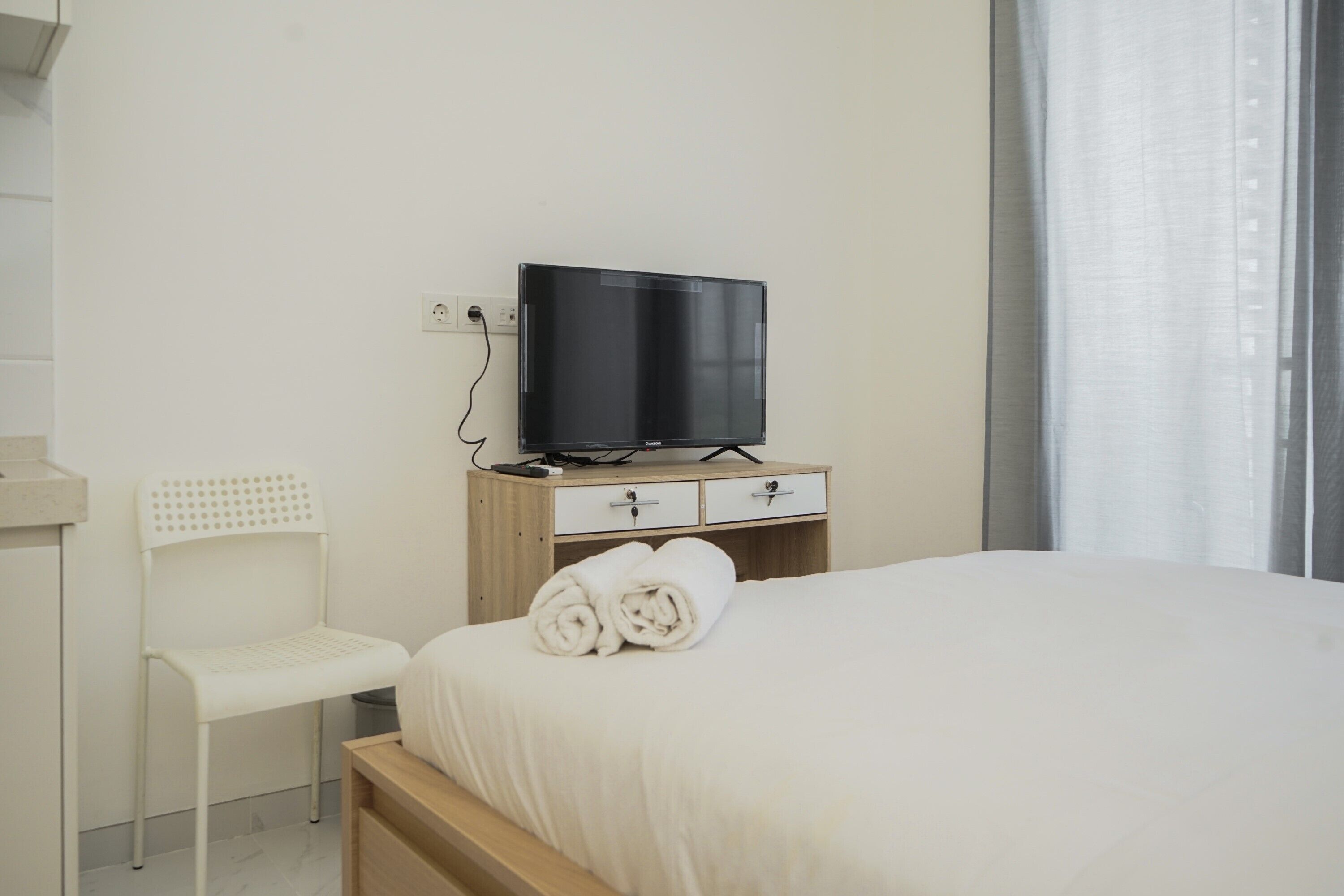 A hotel room with a bed, a TV on a wooden stand, a white chair, and a window with curtains.