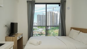 A hotel room with a bed, a TV on a wooden desk, and a view of high-rise buildings through a window. - Gorgeous and Comfy Studio Sky House BSD Apartment (Pagedangan)