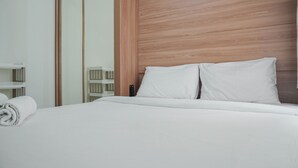A hotel room with a wooden headboard, a neatly made bed with white linens, and a large mirror on the left. - Restful and Tidy 2BR at Green Pramuka City Apartment (Jakarta)