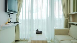 A modern living room with a flat-screen TV on a wall-mounted stand, a sofa, a coffee table, and sheer curtains. - Comfy and Nice 1BR at Tree Park City BSD Apartment (South Tangerang)