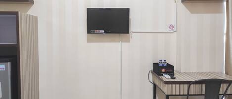 A hotel room with a flat-screen TV mounted on the wall, a desk with a chair, and a refrigerator.