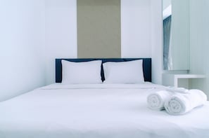 A neatly made bed with white linens and two pillows, a small bedside table with a remote control, and rolled towels on the bed.