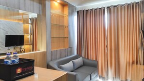 A modern hotel room with a grey sofa, a wooden cabinet, a flat-screen TV, and a tray table with water bottles. - Spacious 2BR at Vida View Makassar Apartment (Makassar)