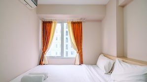 A hotel room with a bed, pillows, a window with curtains, and an air conditioning unit. - Nice and Strategic 2BR at Bassura City Apartment (Jakarta)