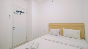 Hotellihuone, jossa on puinen sängynpääty, valkoiset vuodevaatteet, naulakolla varustettu ovi ja lattialla rullalle taitetut pyyhkeet.