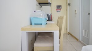A small room with a wooden table and a chair, a blue storage bin, a refrigerator, and a door with a fire alarm.