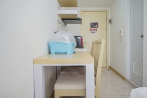 Una habitación pequeña con una mesa de madera y una silla, un contenedor azul para guardar cosas, un refrigerador y una puerta con detector de incendios.