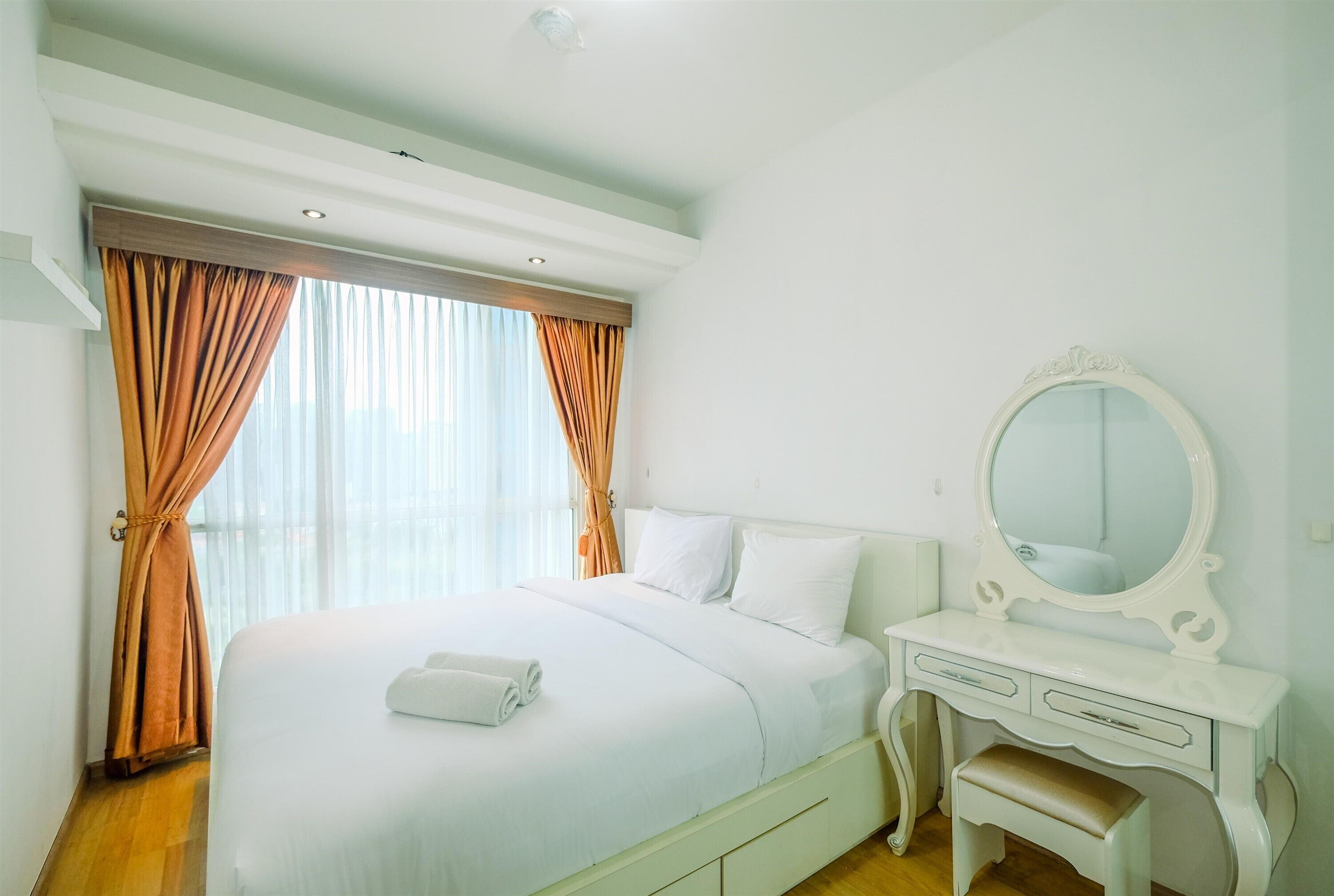 A hotel room with a large bed, white curtains, a vanity with a mirror, and a wooden floor.