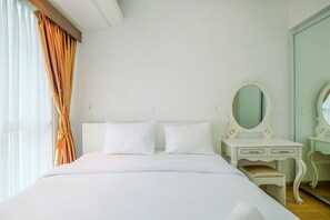 A hotel room with a bed, white curtains, a vanity with a mirror, and a wooden floor.