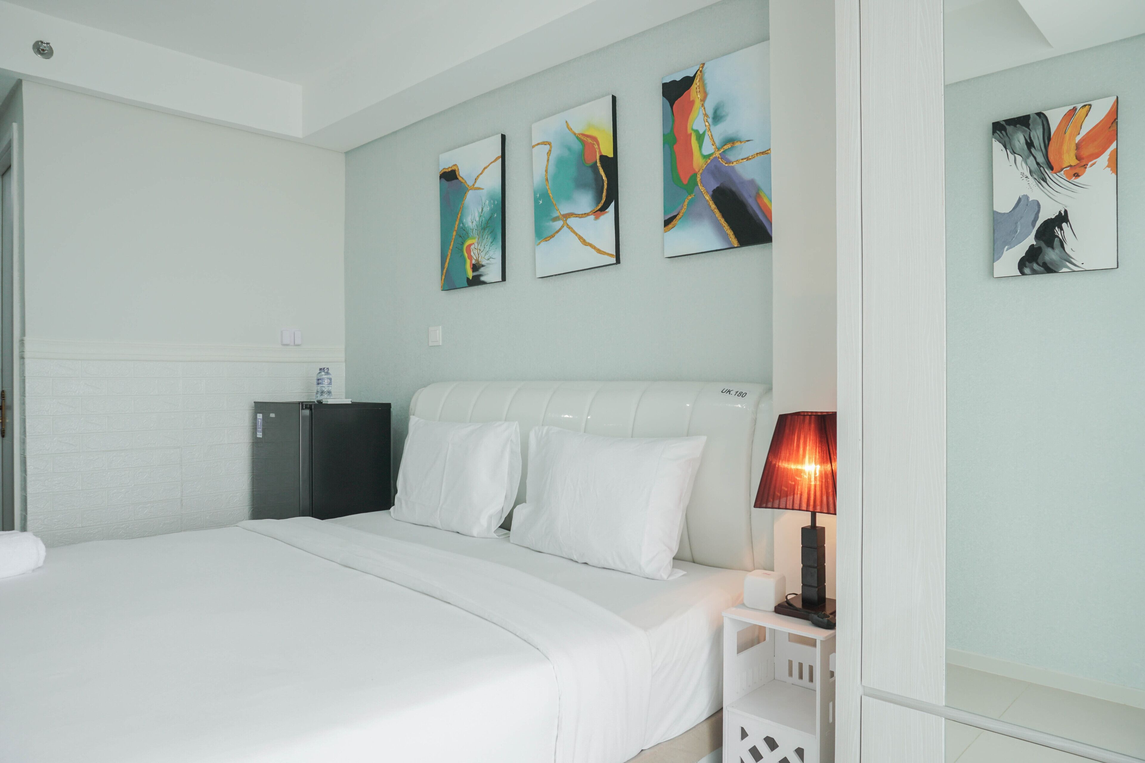 A hotel room with a bed, a nightstand with a lamp, a small cabinet, and three abstract paintings on the wall.