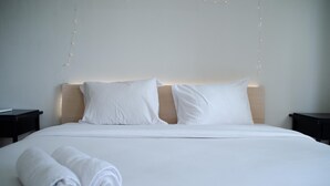 A neatly made bed with white linens and pillows, two rolled white towels, and string lights on the wall.