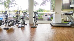 A gym with stationary bikes and a treadmill, large windows, and a view of parked cars and trees outside.