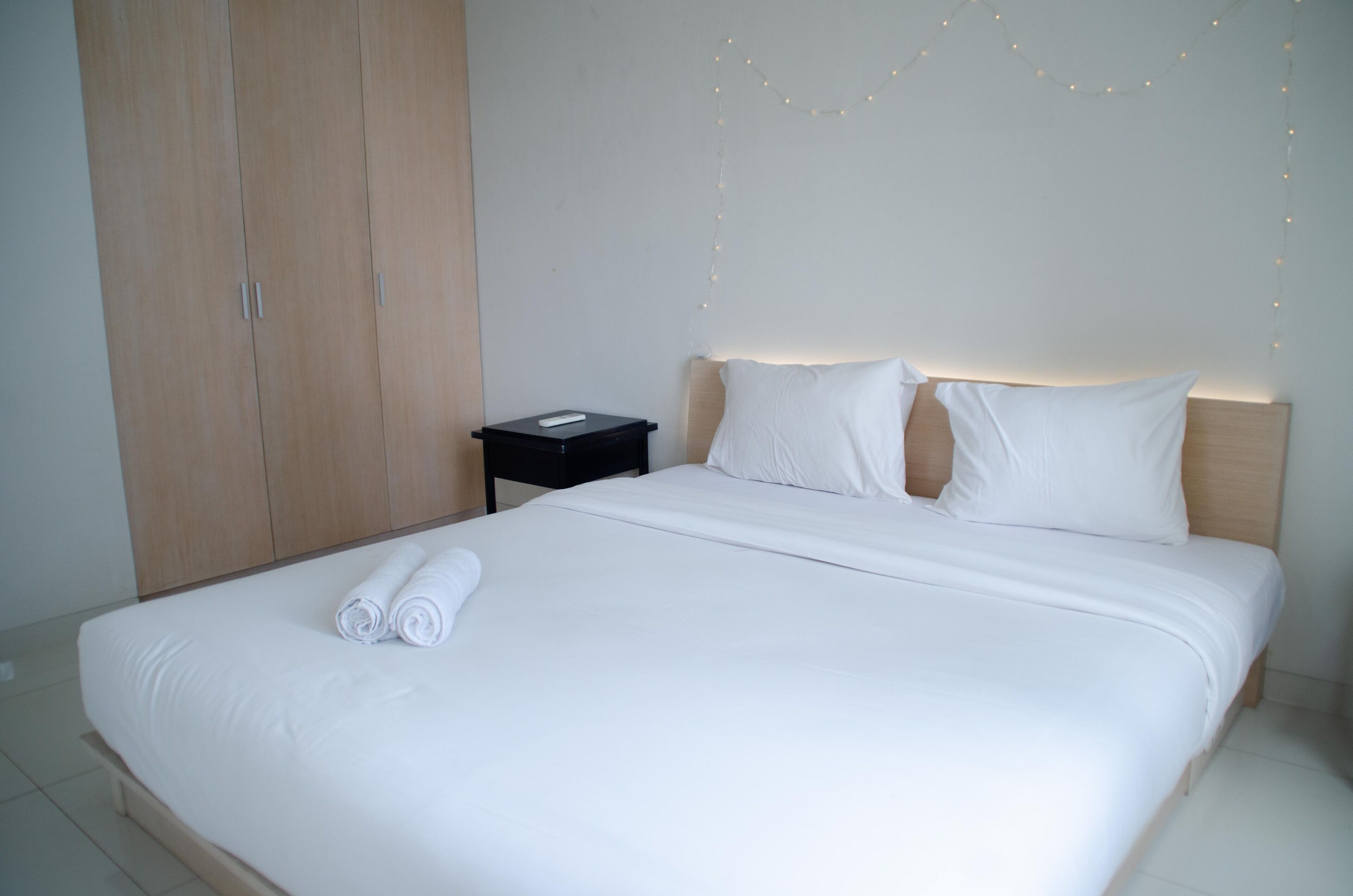 Una cama bien tendida con sábanas blancas y toallas enrolladas, una mesita de noche con lámpara y un armario.