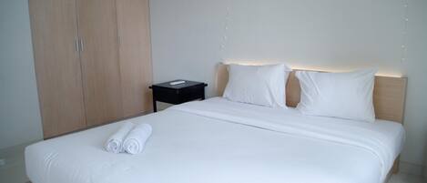 Una cama bien tendida con sábanas blancas y toallas enrolladas, una mesita de noche con lámpara y un armario.