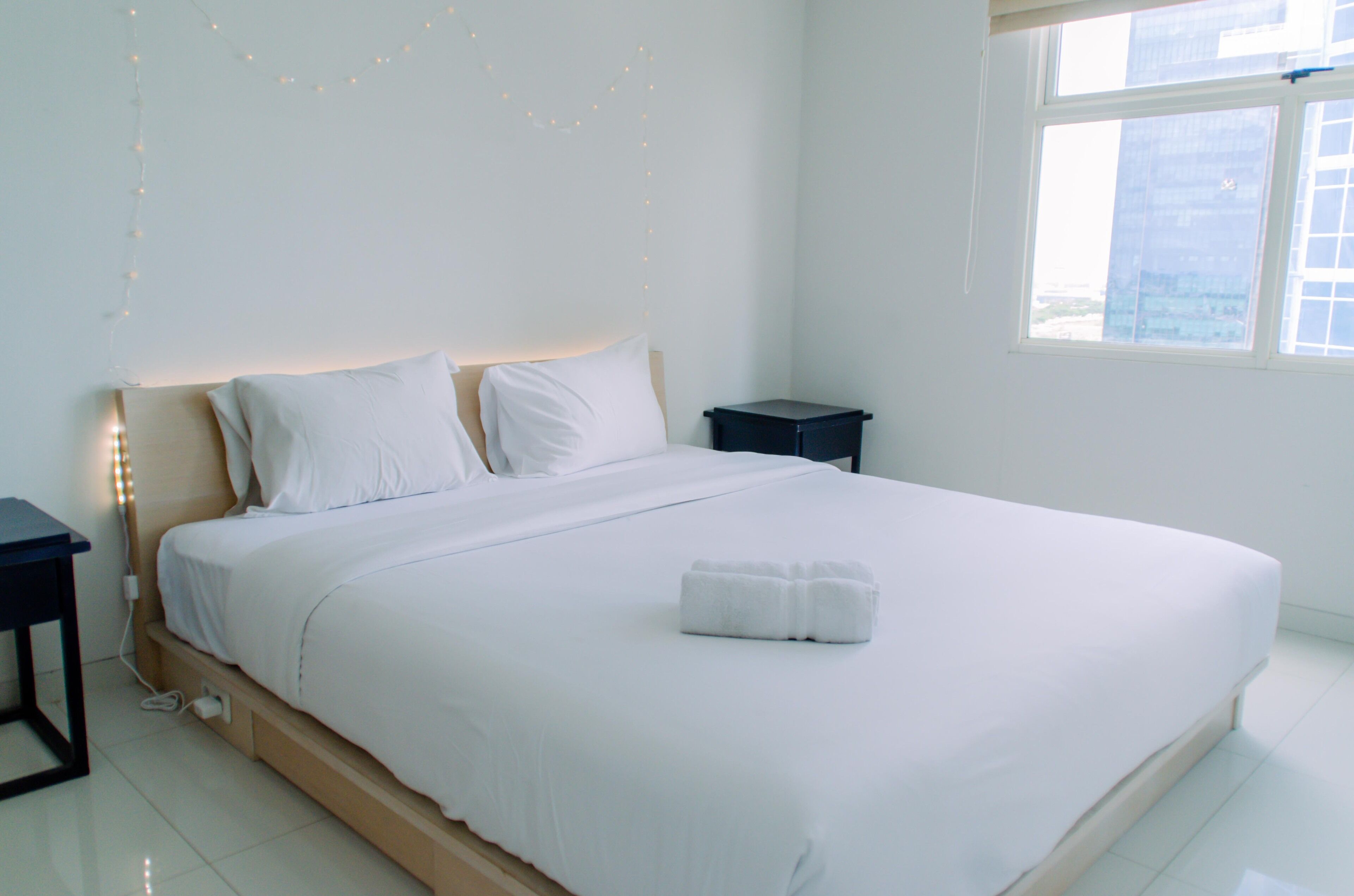 A neatly made bed with white linens and a folded towel, a bedside table, and a nightstand with a lamp.
