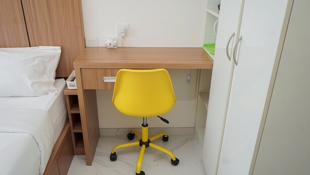 Une petite chambre à coucher, bien rangée, avec un bureau en bois, une chaise de bureau jaune, un lit blanc et une armoire blanche.