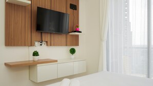 Una habitación de hotel moderna con un televisor de pantalla plana montado en una pared de madera, un estante de madera con una planta pequeña, una mesita de noche blanca con toallas enrolladas y una ventana con cortinas traslúcidas.