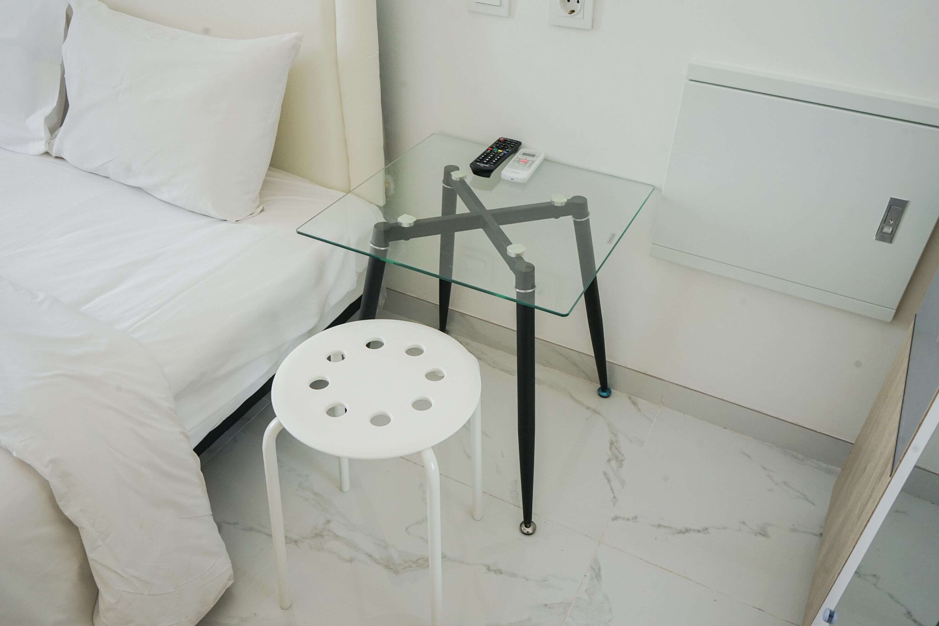 A room with a bed, a glass table with a remote control, and a white chair.