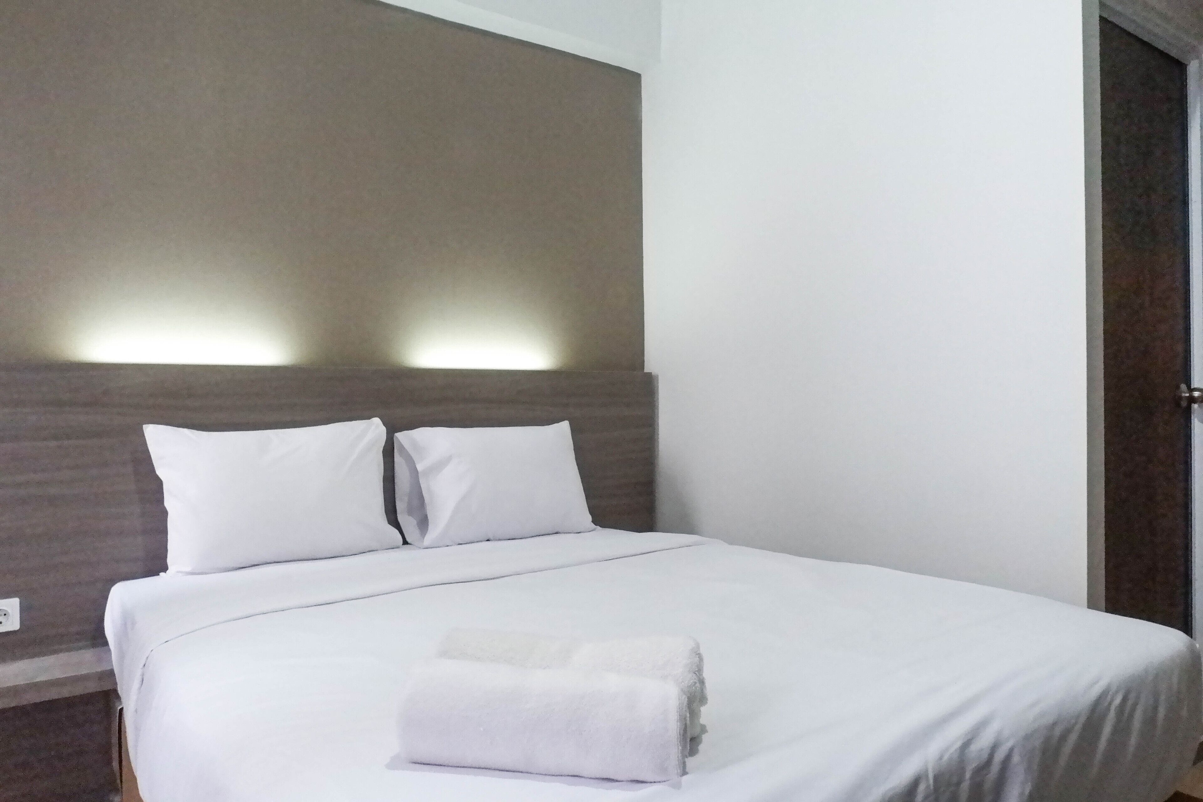 Un lit bien fait, avec du linge de lit blanc et des oreillers, une serviette blanche pliée posée sur le lit, et un luminaire fixé au mur au-dessus.