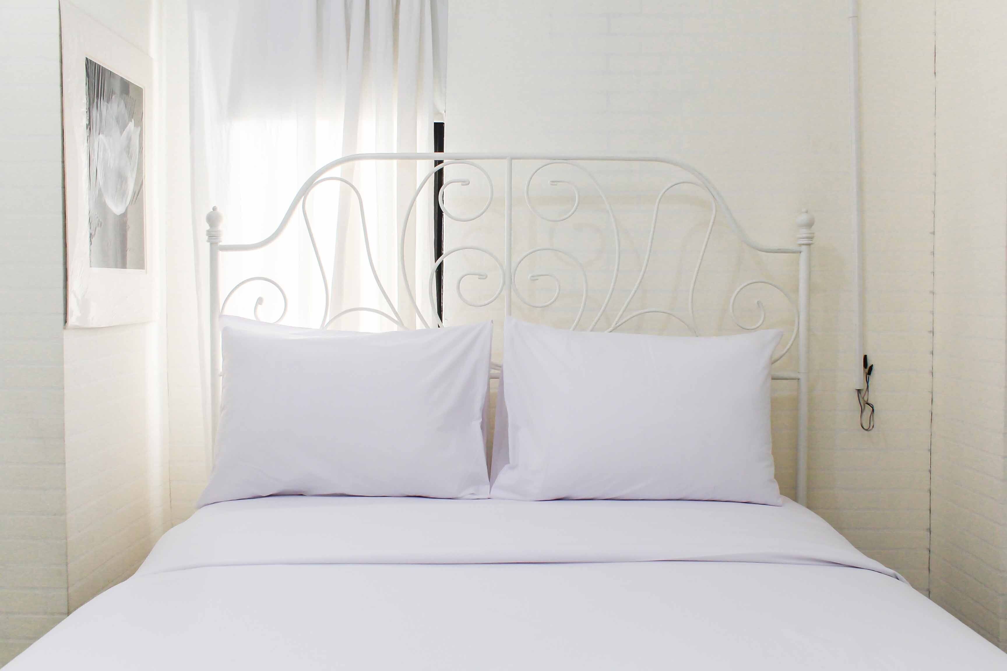 Estructura de cama metálica blanca con dos almohadas y una colcha blanca en una habitación con paredes de ladrillo blanco y una ventana con cortinas translúcidas.