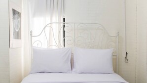 A white metal bed frame with two pillows and a white bedspread in a room with white brick walls and a window with sheer curtains. - White and Nice 1BR at Bassura City Apartment (Jakarta)