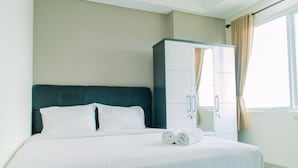A neatly made bed with white linens and rolled towels, a dark headboard, a wardrobe with two doors, and a window with curtains. - Minimalist and Cozy Living Studio Apartment at B Residence (Pagedangan)