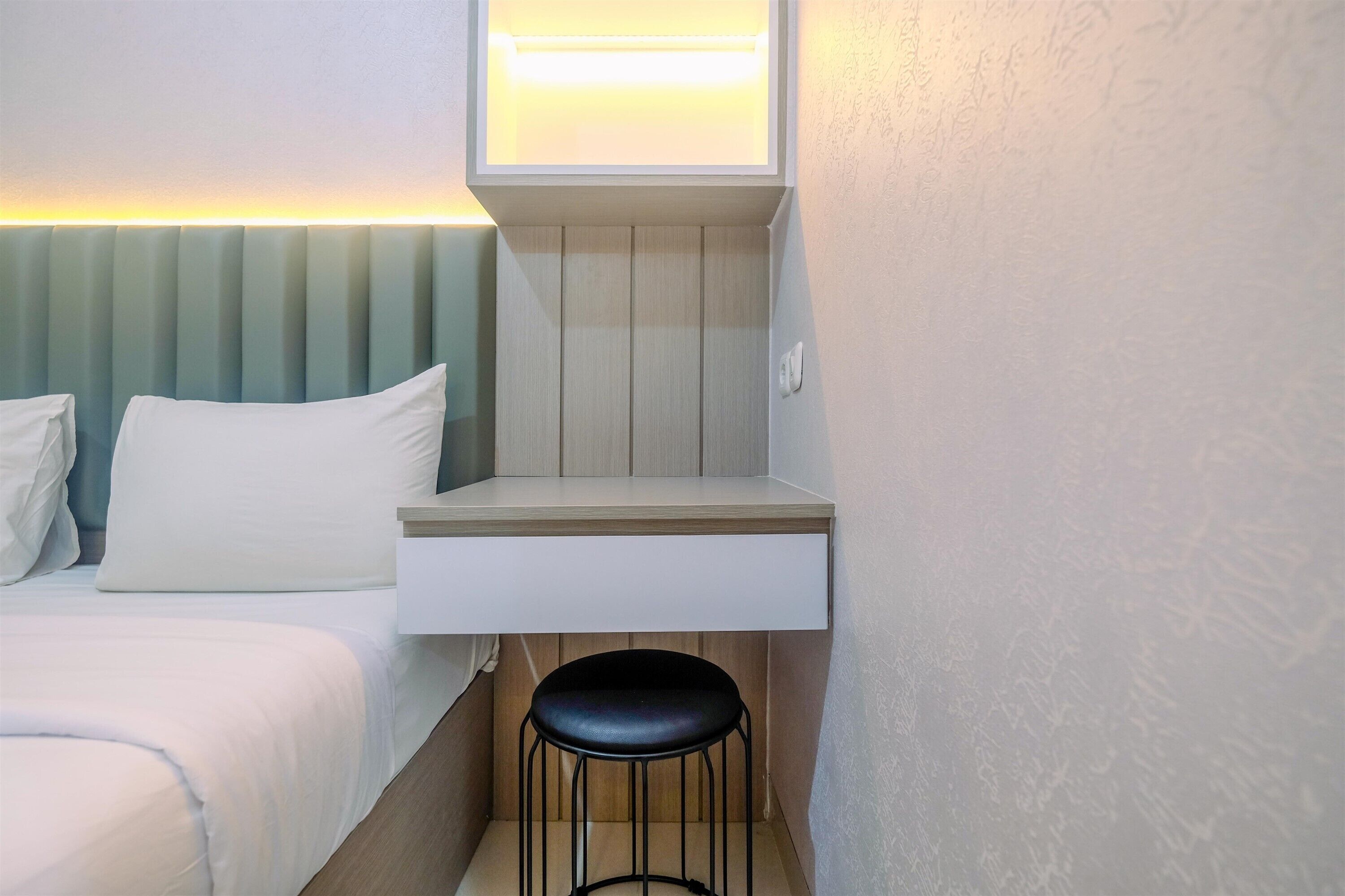Une chambre d’hôtel de taille réduite, comprenant un lit, un petit bureau avec une chaise et un luminaire fixé au mur.