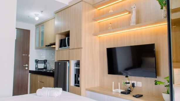 Une chambre d’hôtel moderne équipée d’un téléviseur à écran plat fixé au mur, d’un lit avec du linge de maison blanc, d’un petit coin cuisine avec un réfrigérateur et un four à micro-ondes, et d’une plante en pot sur la table de chevet.