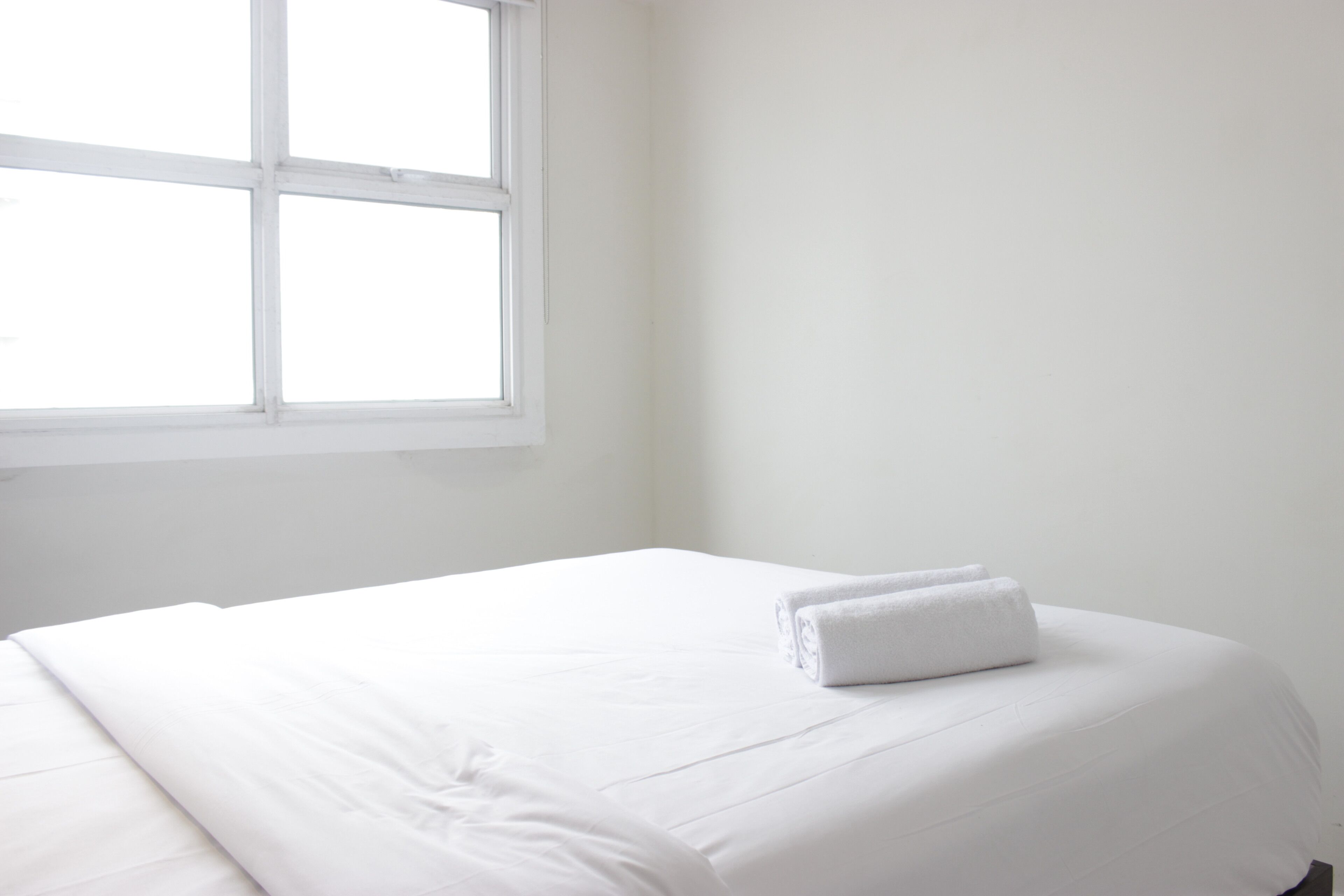 A neatly made bed with white linens and a rolled towel on the side, a window with white frames, and a plain white wall.