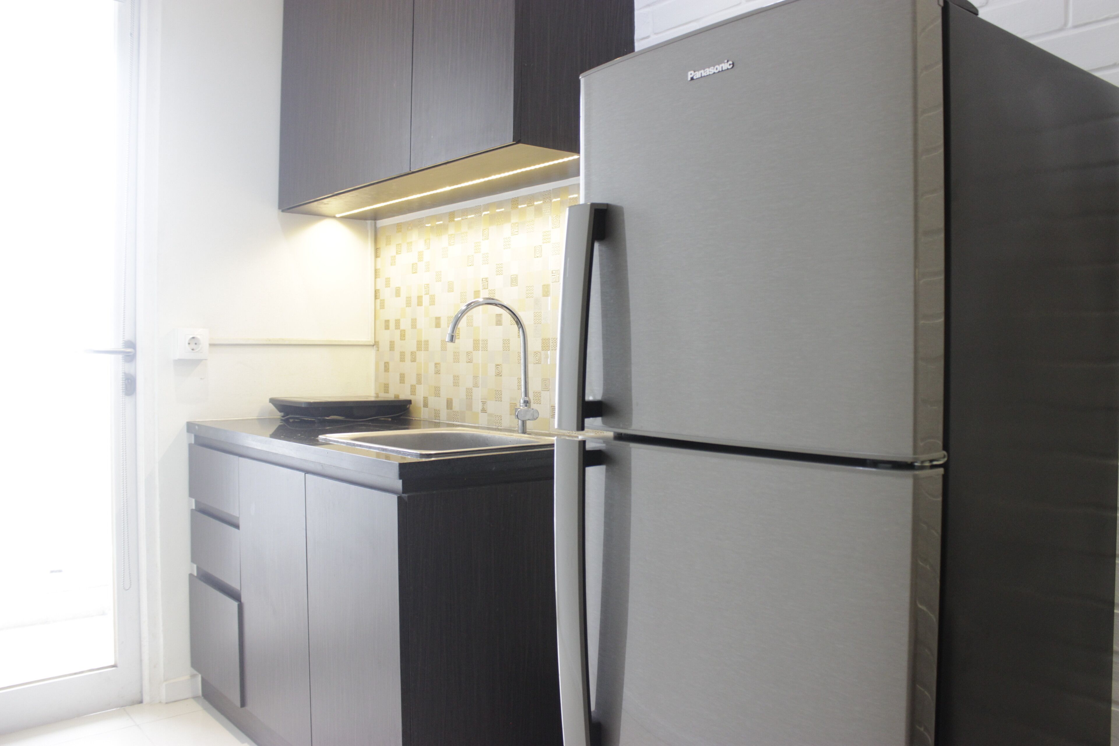 A kitchen with a stainless steel refrigerator, dark wooden cabinets, a tiled backsplash, and a window providing natural light.