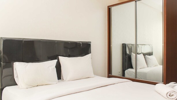 A neatly made bed with white linens and pillows, a large mirror reflecting the room, and a wooden wardrobe. - Nice and Elegant Studio at Riverview Jababeka Apartment (Cikarang)