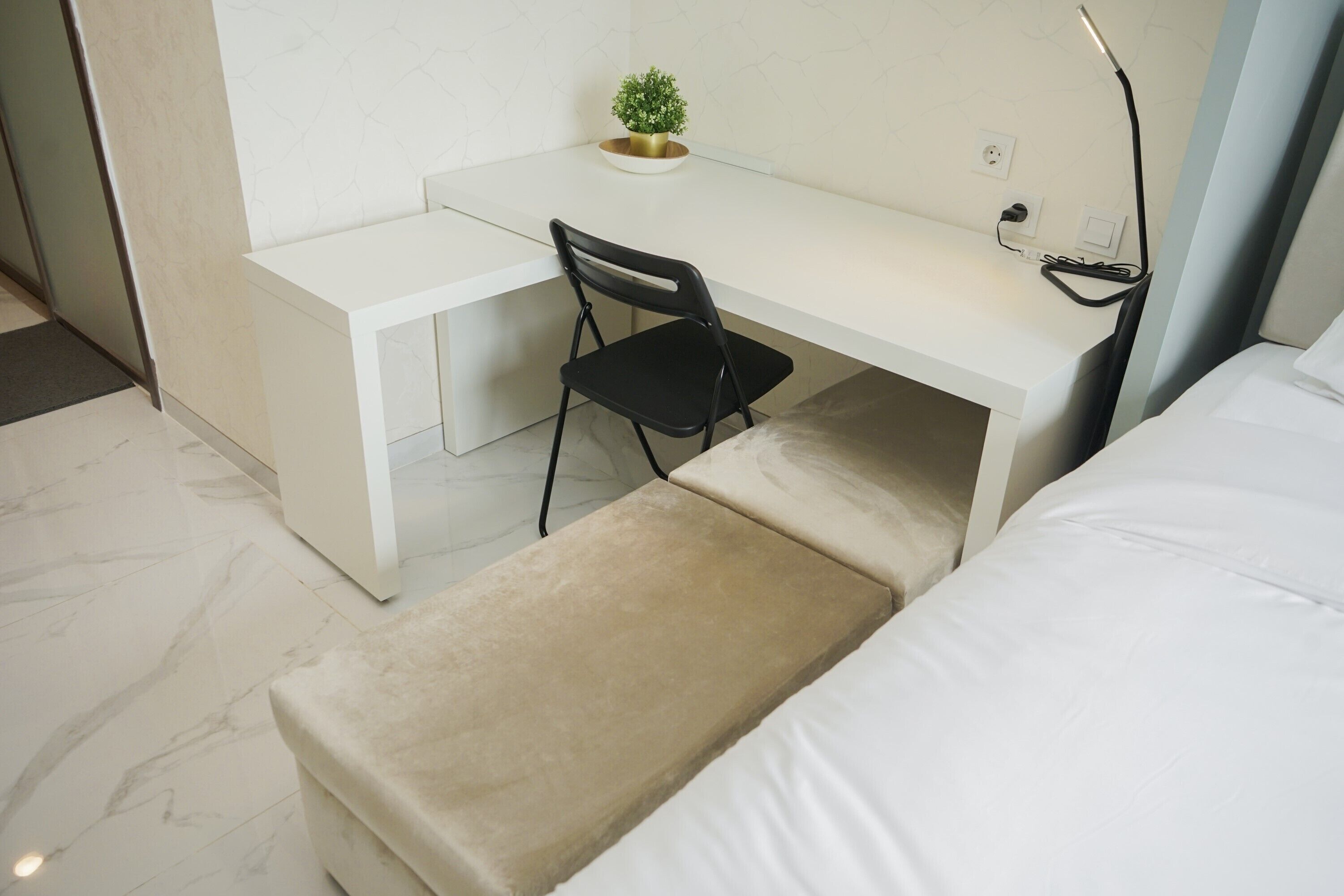 A modern hotel room with a white desk, a black chair, a small potted plant, and a bed with white linens.