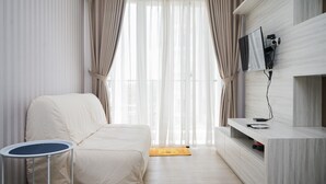 A modern hotel room with a large window, a beige sofa, a small round table, and a mounted flat-screen TV.