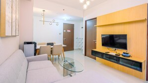 Ett modernt vardagsrum med en soffa, ett soffbord i glas och en platt-TV som är monterad på en väggpanel i trä. I bakgrunden finns en matplats med ett bord och stolar.