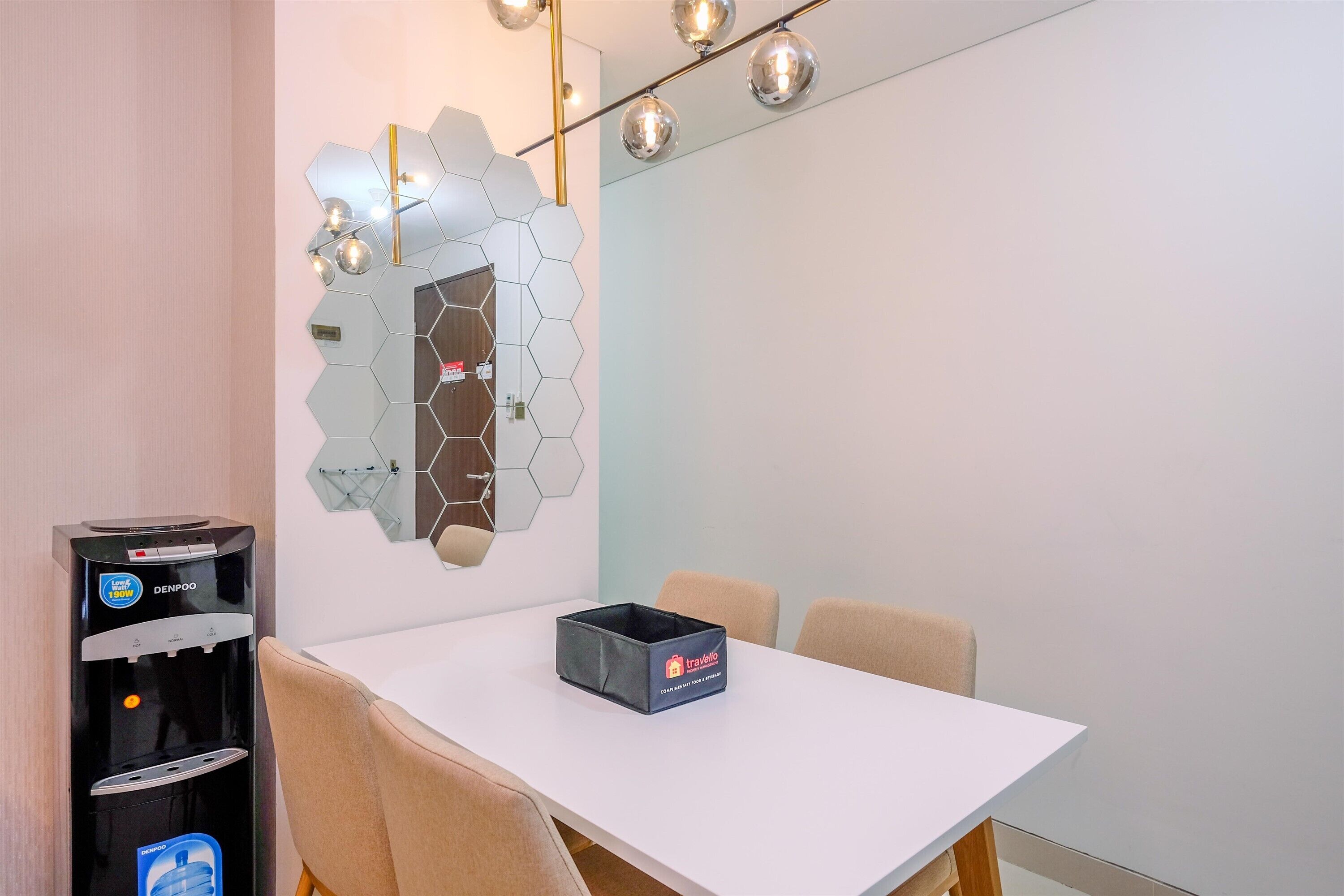 Quarto de hotel moderno com uma mesa branca, cadeiras bege, dispensador de água e um espelho hexagonal decorativo na parede.