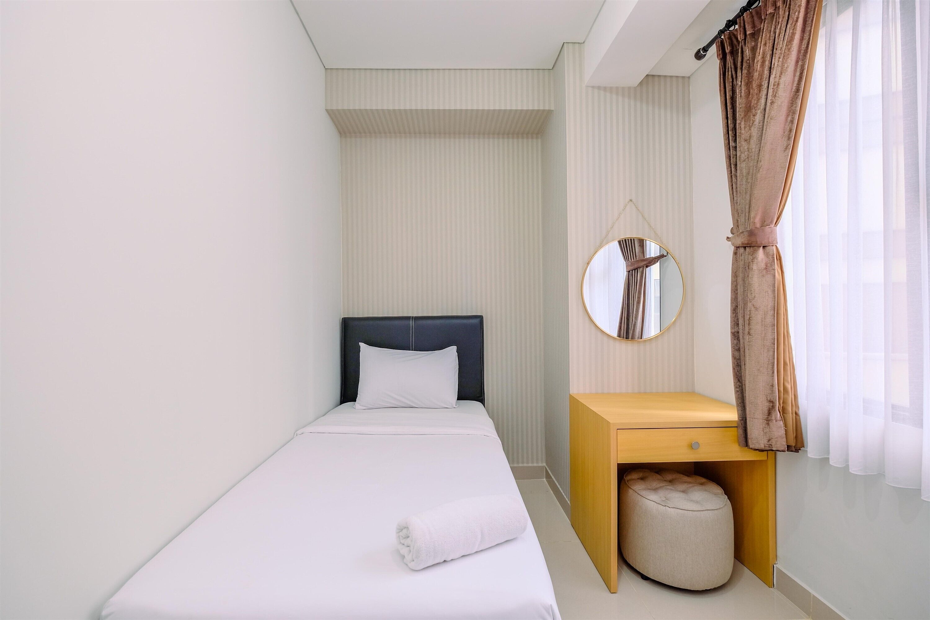 Een netjes ingerichte slaapkamer met een eenpersoonsbed, nachtkastje, ronde spiegel en een raam met gordijnen.