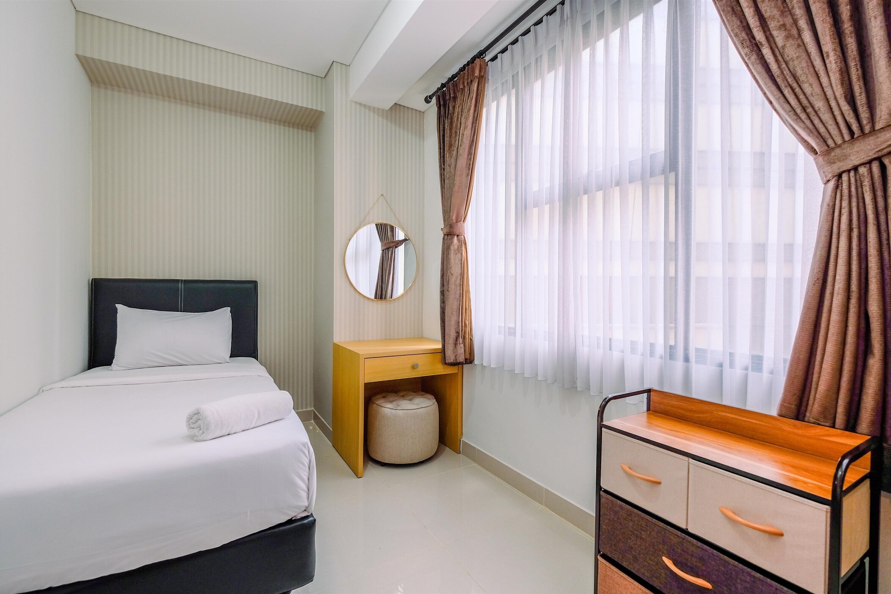 Una habitación de hotel con una cama, una mesita de noche con espejo, una mesita auxiliar de madera, un pequeño reposapiés y un aparador con cajones.