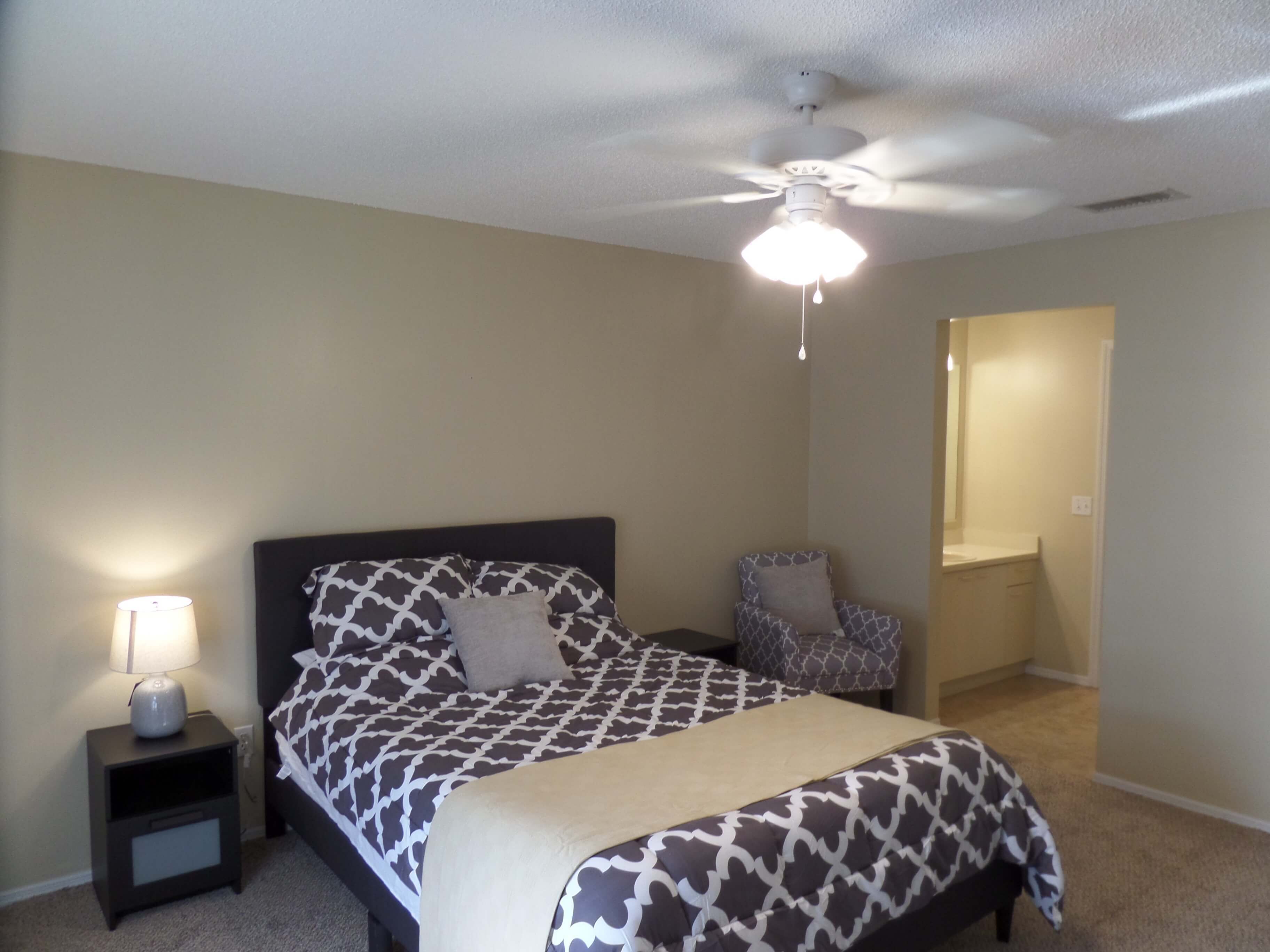 A bedroom with a bed, a nightstand, a chair, a ceiling fan, and a bathroom door.