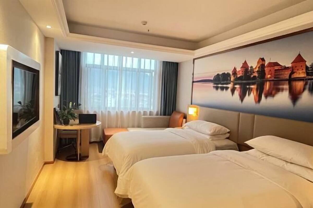Habitación de hotel con dos camas, un escritorio con una computadora portátil, una ventana con cortinas y un cuadro de un castillo en la pared.