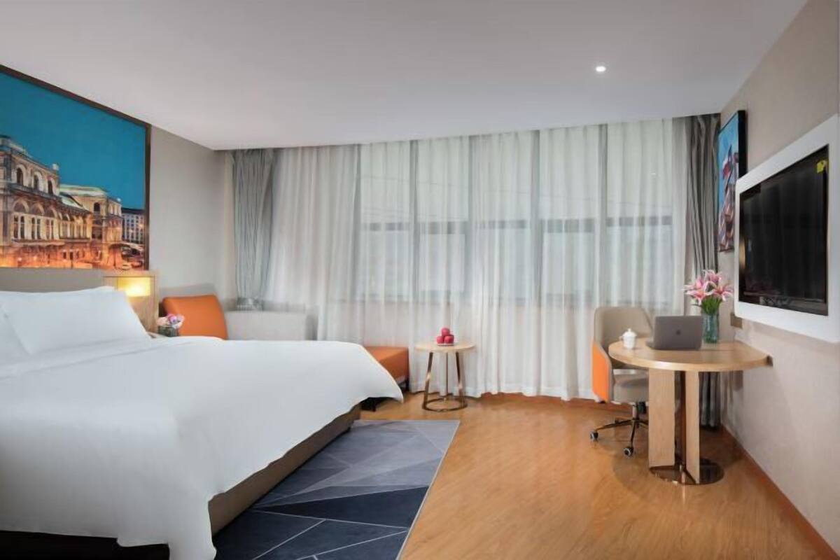 Habitación de hotel con una cama grande, un escritorio con una computadora portátil, una silla, una mesita y un ventanal con cortinas.