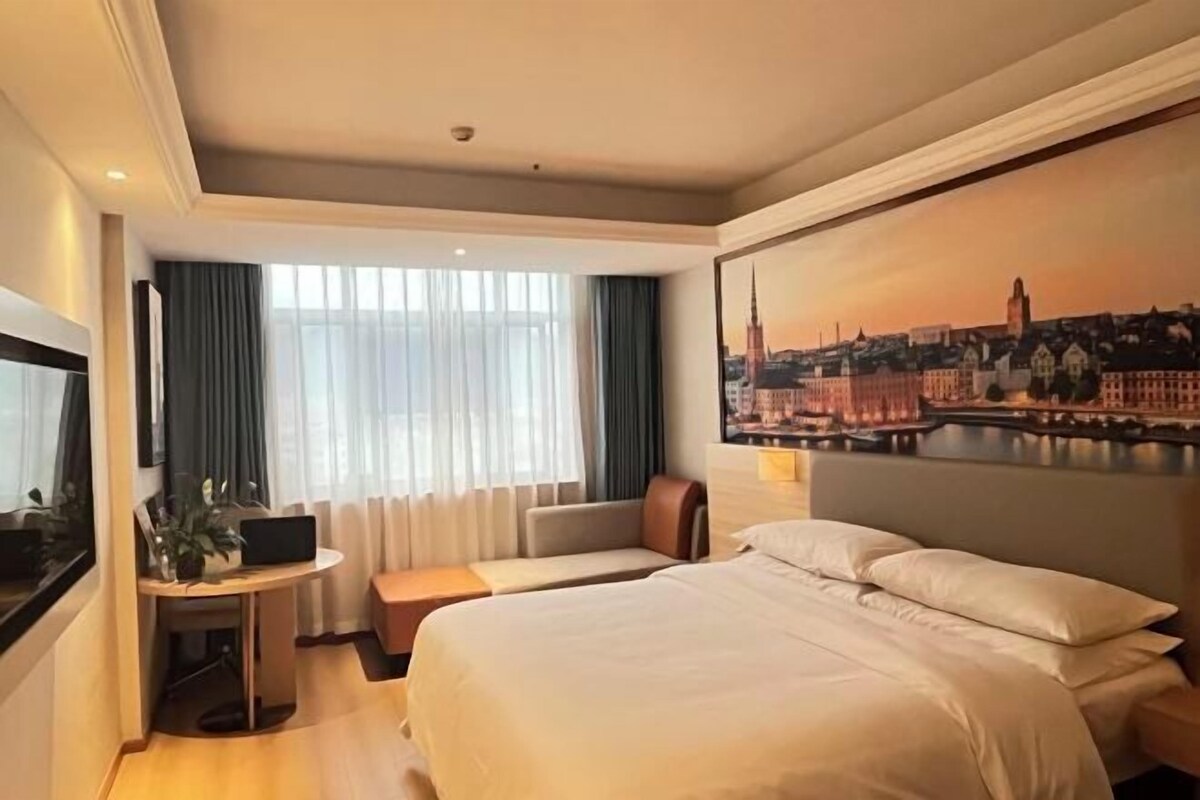 A hotel room with a large bed, a desk with a laptop, a seating area, and a cityscape painting on the wall.