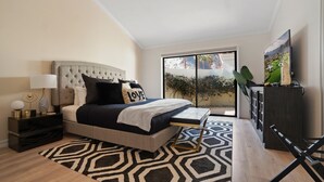 A modern bedroom with a tufted headboard, a large bed with a geometric patterned rug, a bedside table with a lamp, a TV on a stand, and a view of a landscaped area through a sliding door.